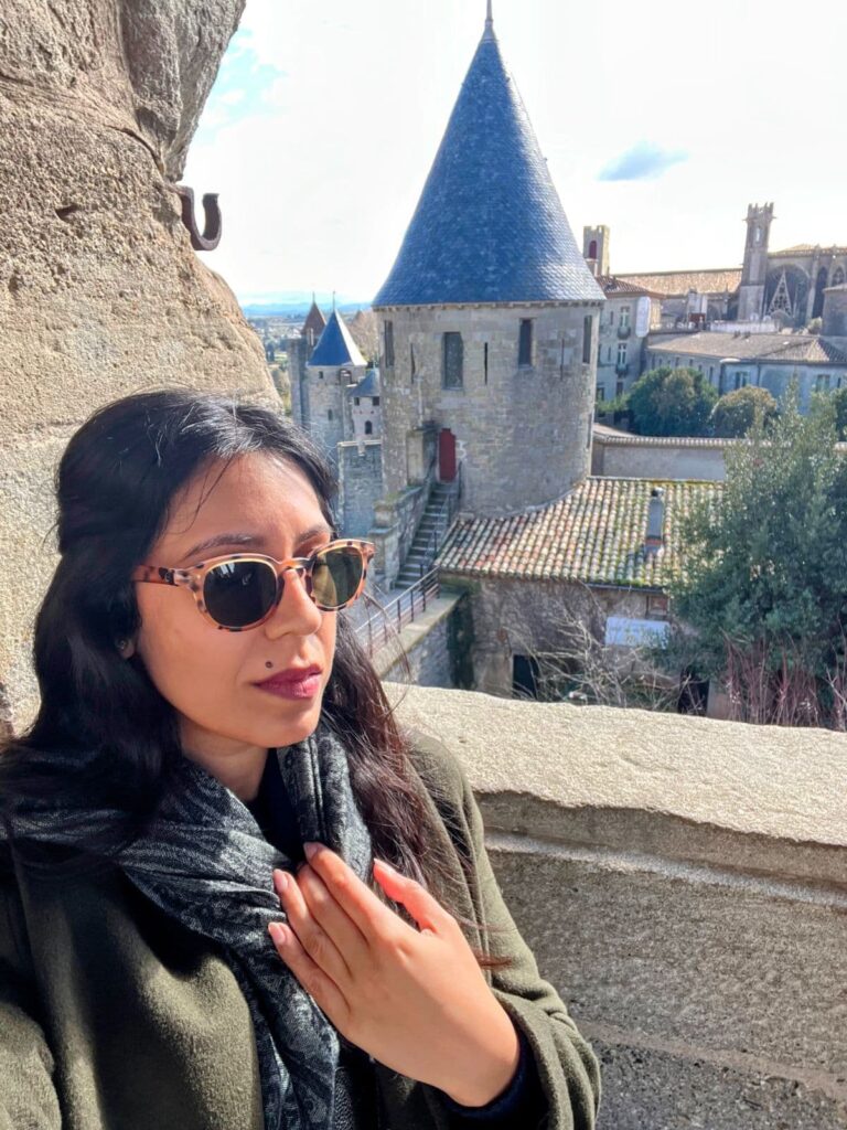A woman in sunglasses stands near a stone window overlooking the medieval towers and rooftops of Carcassonne, France, with blue spires and historic architecture in view.