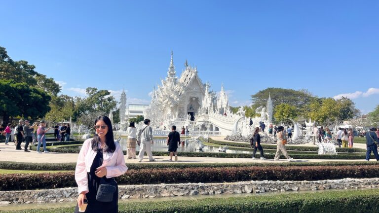Traveling alone as a woman, she smiles in front of an ornate white temple with intricate carvings and mirrored details under a bright blue sky. Visitors walk around the reflective pool and landscaped gardens, highlighting the temple as a popular travel destination.