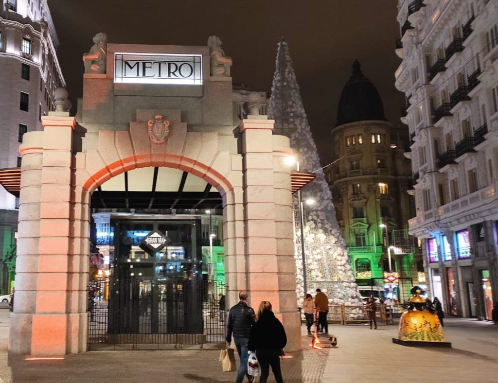 A Metro entrance stands illuminated at night, framed by historic buildings and festive lights. A large decorated Christmas tree rises nearby while pedestrians walk past the station entrance.