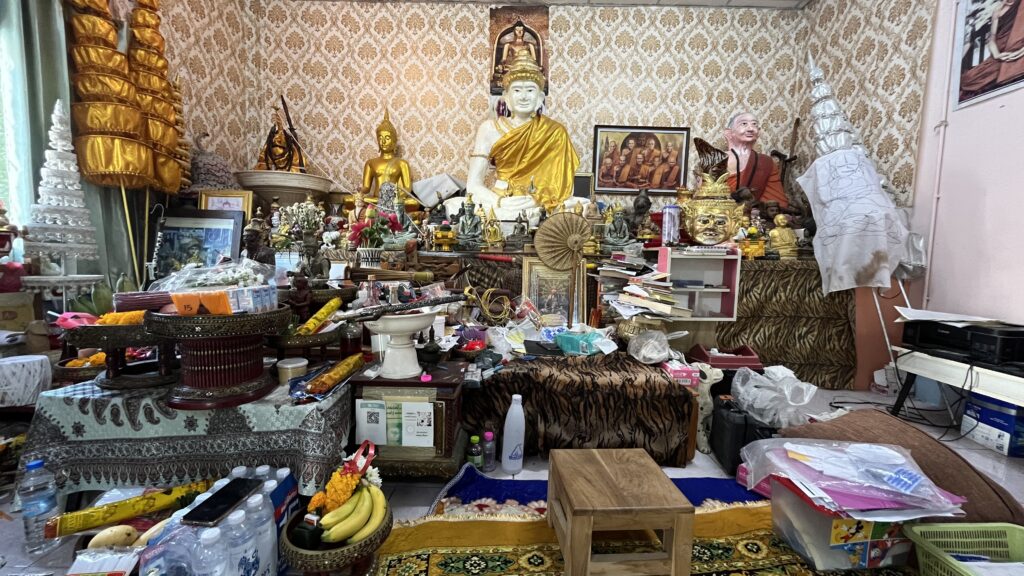 Shrine room filled with Buddha statues monk portraits ritual masks offerings incense and ceremonial objects arranged across crowded tables behind a patterned prayer mat and small wooden stool.