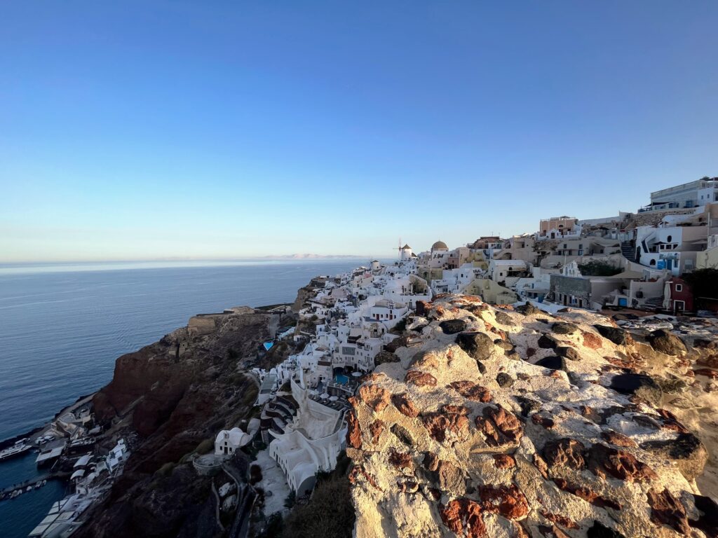 Panoramic view of Oia Santorini with white cave houses stacked along the caldera cliffs above the deep blue Aegean Sea at golden hour.