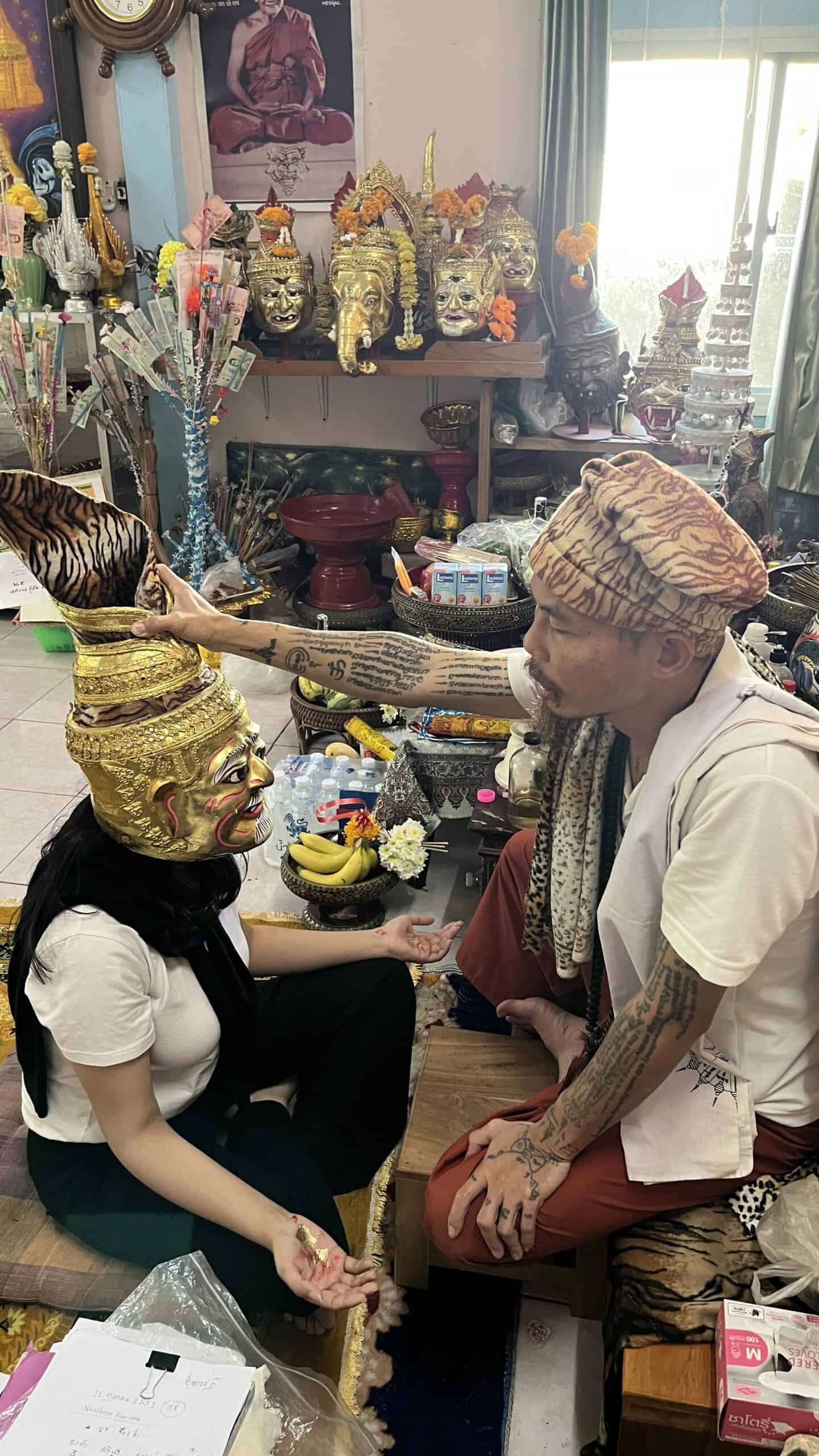Spiritual practitioner adjusts a large golden ritual mask with a tall tiger patterned headdress on a seated woman inside a shrine room filled with statues offerings and flowers.