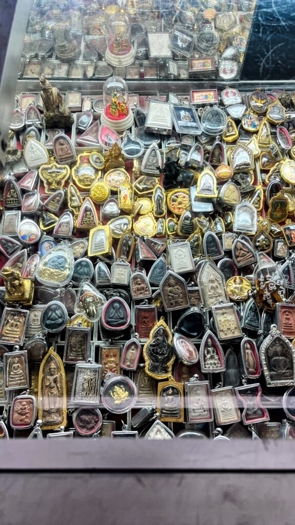 Glass display case packed tightly with dozens of Buddhist amulets and pendants in different shapes colors and metal frames arranged in rows.
