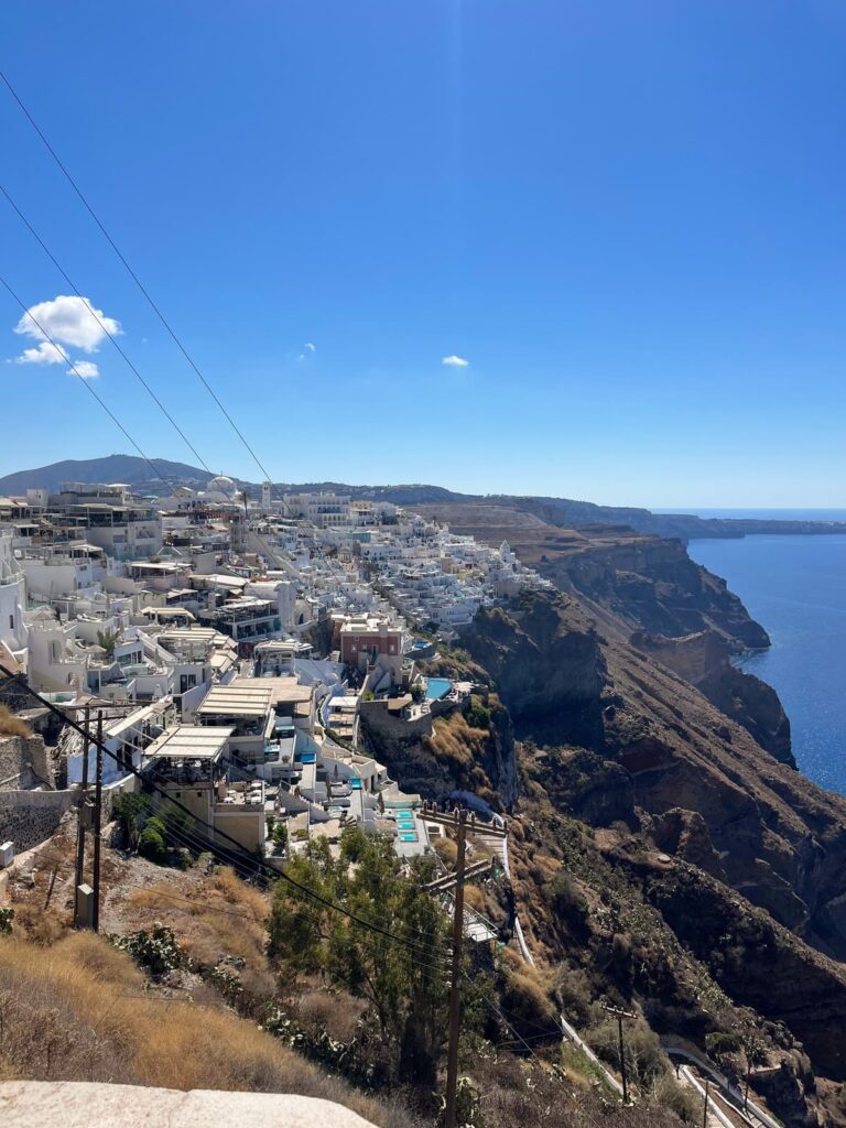 White cliffside hotels and homes line the edge of the Santorini caldera under a bright blue sky with the deep blue Aegean Sea stretching beyond. The elevated viewpoint captures the dramatic drop to the water and layered island landscape.