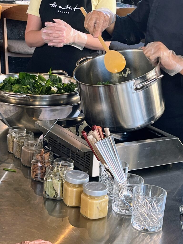 Two people wearing gloves cooking greens in a large pot on a portable stove surrounded by jars of spices and ingredients during an Athens cooking class.