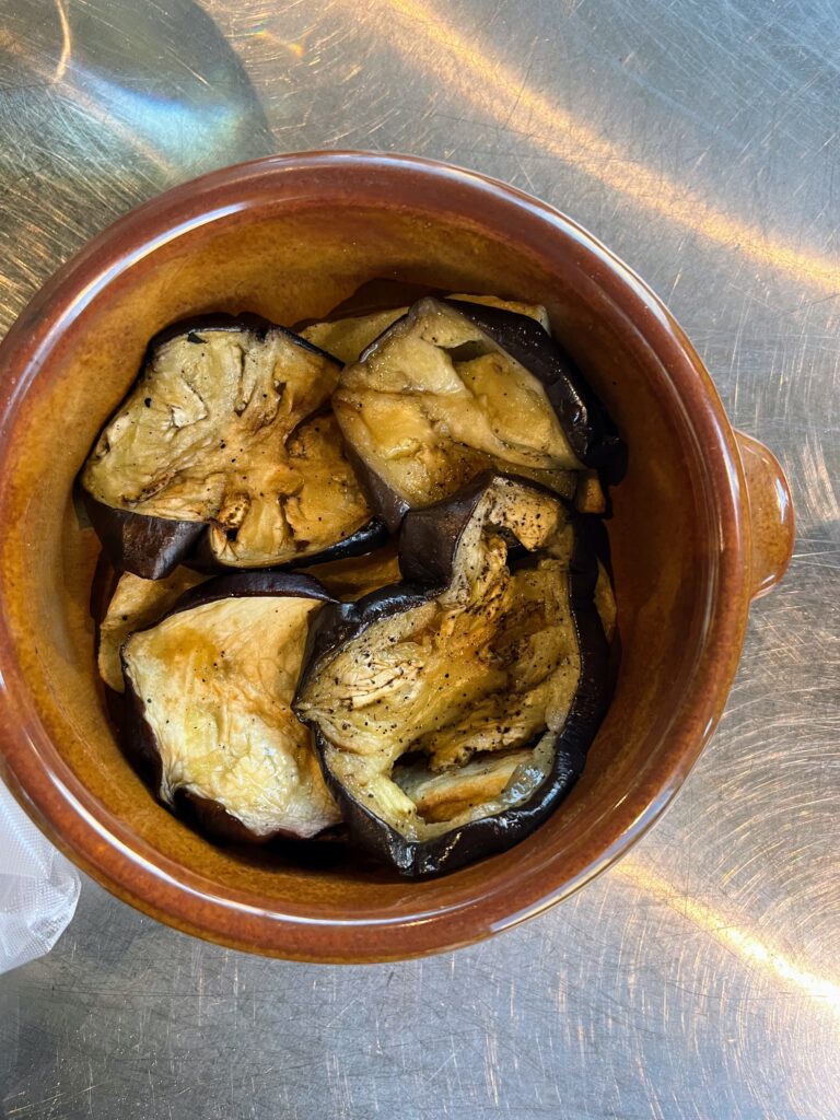 Roasted eggplant slices seasoned and served in a brown ceramic dish on a stainless steel surface.