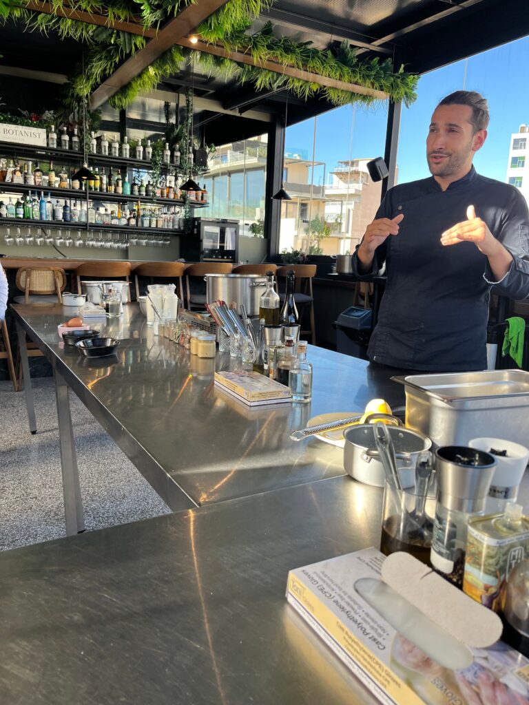 Chef in a black uniform explaining cooking techniques to a group around a stainless steel prep table during an Athens cooking class.