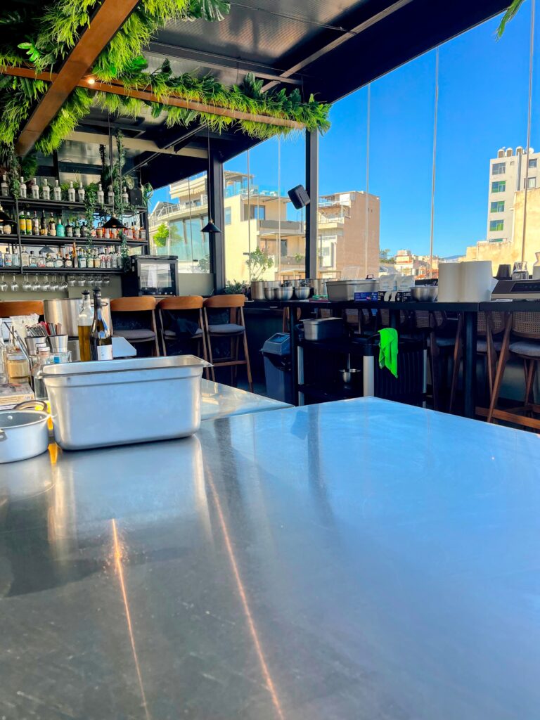 Bright open kitchen with stainless steel counters bar seating and hanging greenery overlooking city buildings through large windows in Athens.