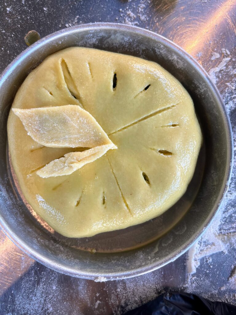 Unbaked pie with smooth dough scored on top and decorative pastry leaves placed in a round metal pan on a floured surface, part of Athens cooking class