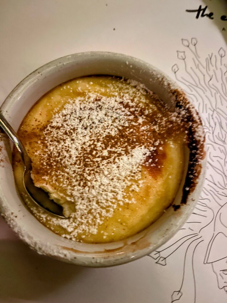 Small ramekin of creamy dessert dusted with powdered sugar and cinnamon with a spoon breaking into the soft center on a plate.