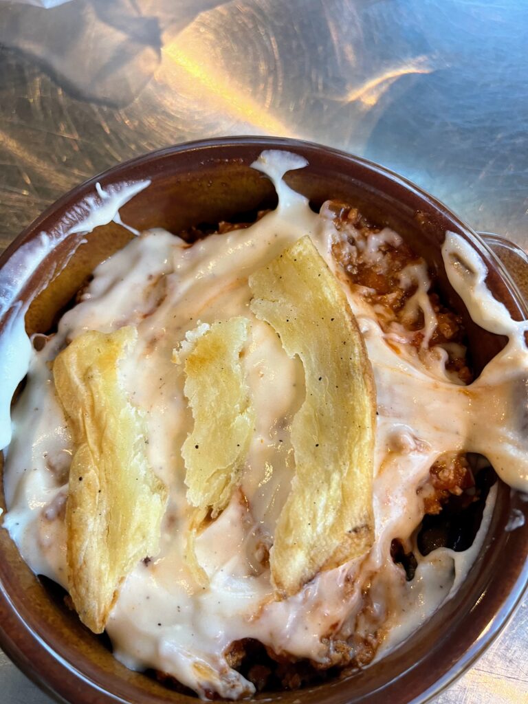 Close up of a baked dish with melted cheese and layers of meat sauce topped with crispy potato slices in a small clay pot during a cooking class.