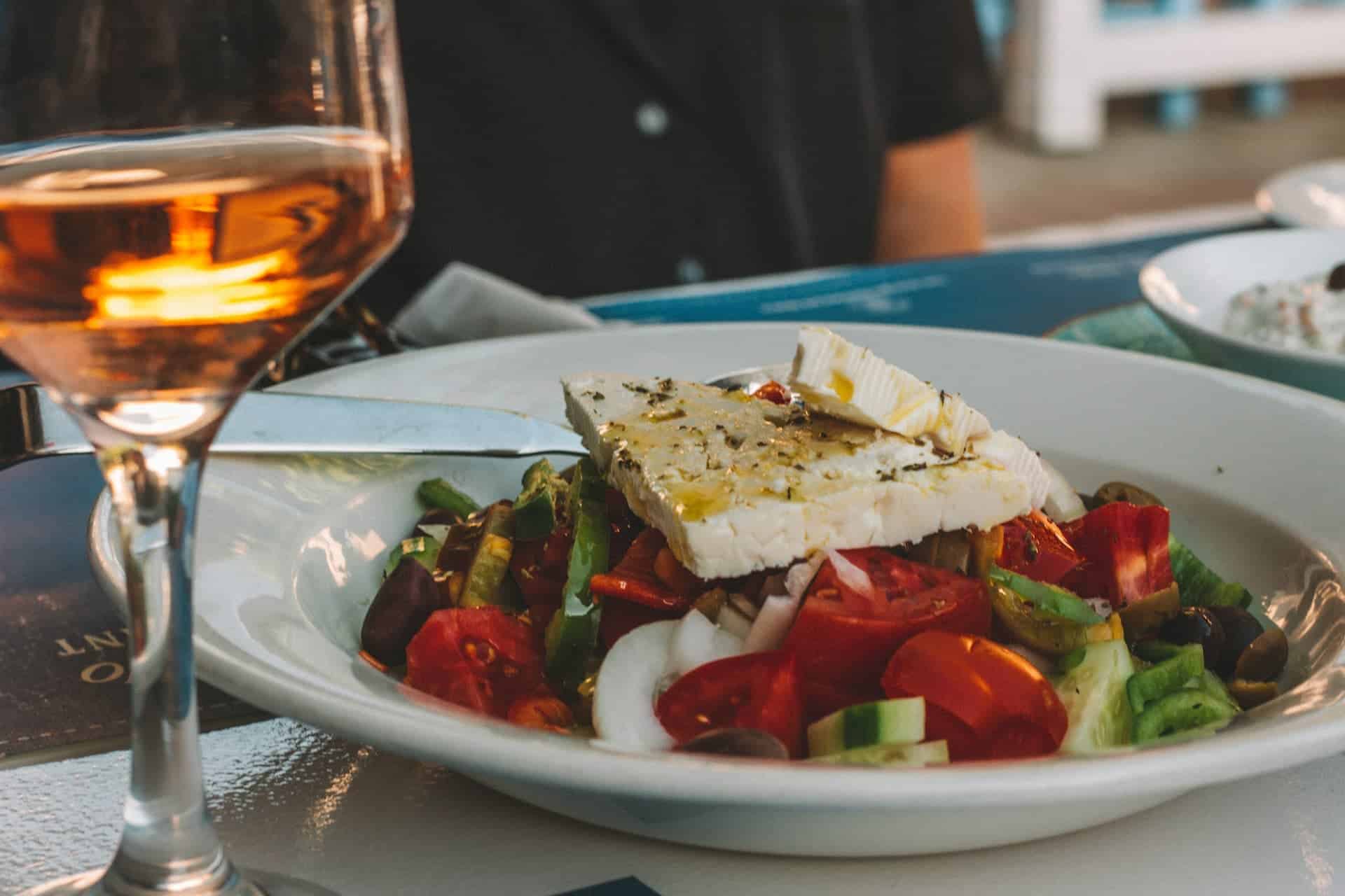 Greek salad topped with a thick slice of feta cheese and olive oil served on a white plate beside a glass of rosé wine at a restaurant table.