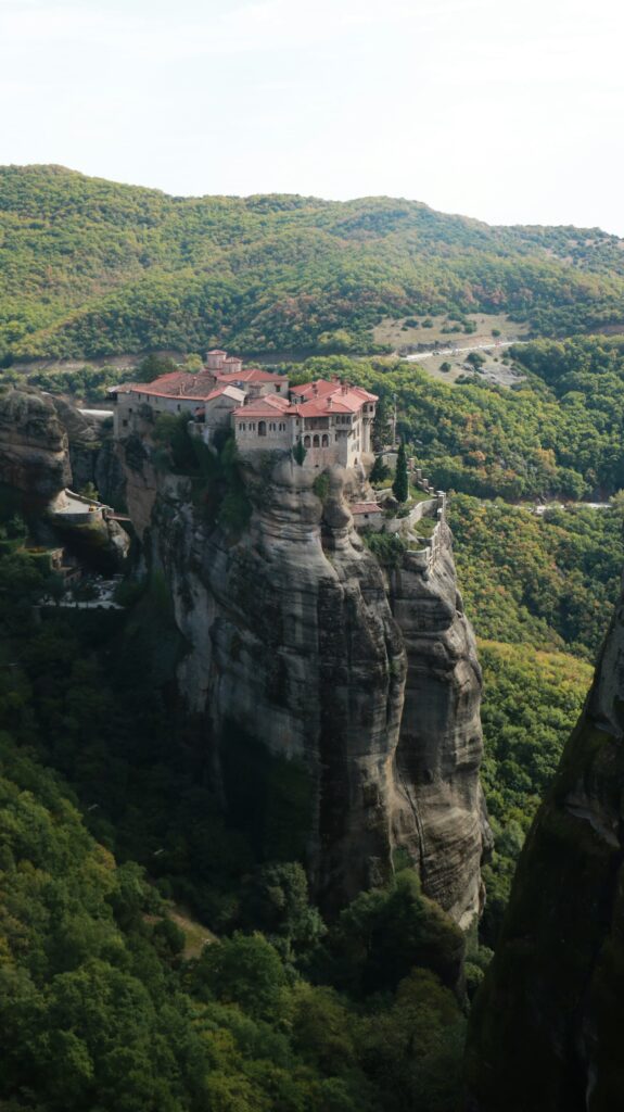 Monastery with red tiled roofs perched dramatically on top of a tall rock formation surrounded by lush green landscape in Meteora.