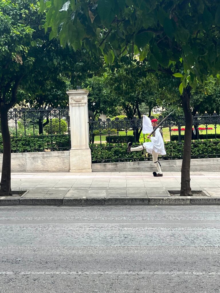Evzones guard in traditional uniform marching with high step along a tree lined street in Athens with an iron fence and garden behind him.