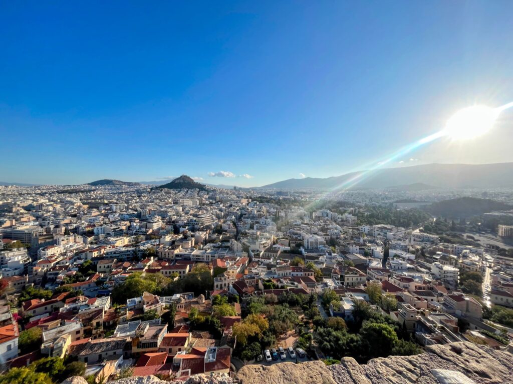 Panoramic view over Athens with white buildings stretching across the city and hills in the distance under a bright sun in a clear blue sky.