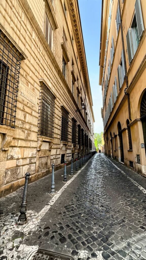 Empty cobblestone alley between historic stone buildings in Rome with iron window grilles and soft afternoon light at the end of the street.