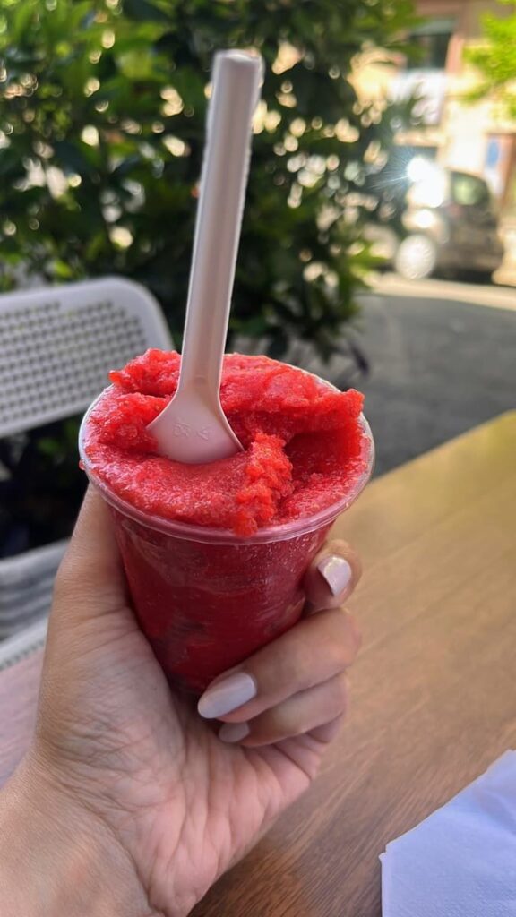 Hand holding a bright red frozen granita in a clear plastic cup at an outdoor café in Italy. Green plants and a parked car blur into the background on a sunny afternoon.