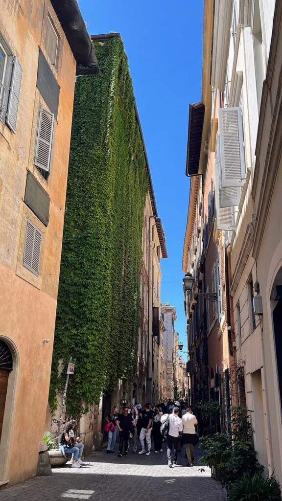 cNarrow street in Rome with people walking past a tall building completely covered in green ivy. Sunlight reflects off the pastel colored buildings beneath a clear blue sky.