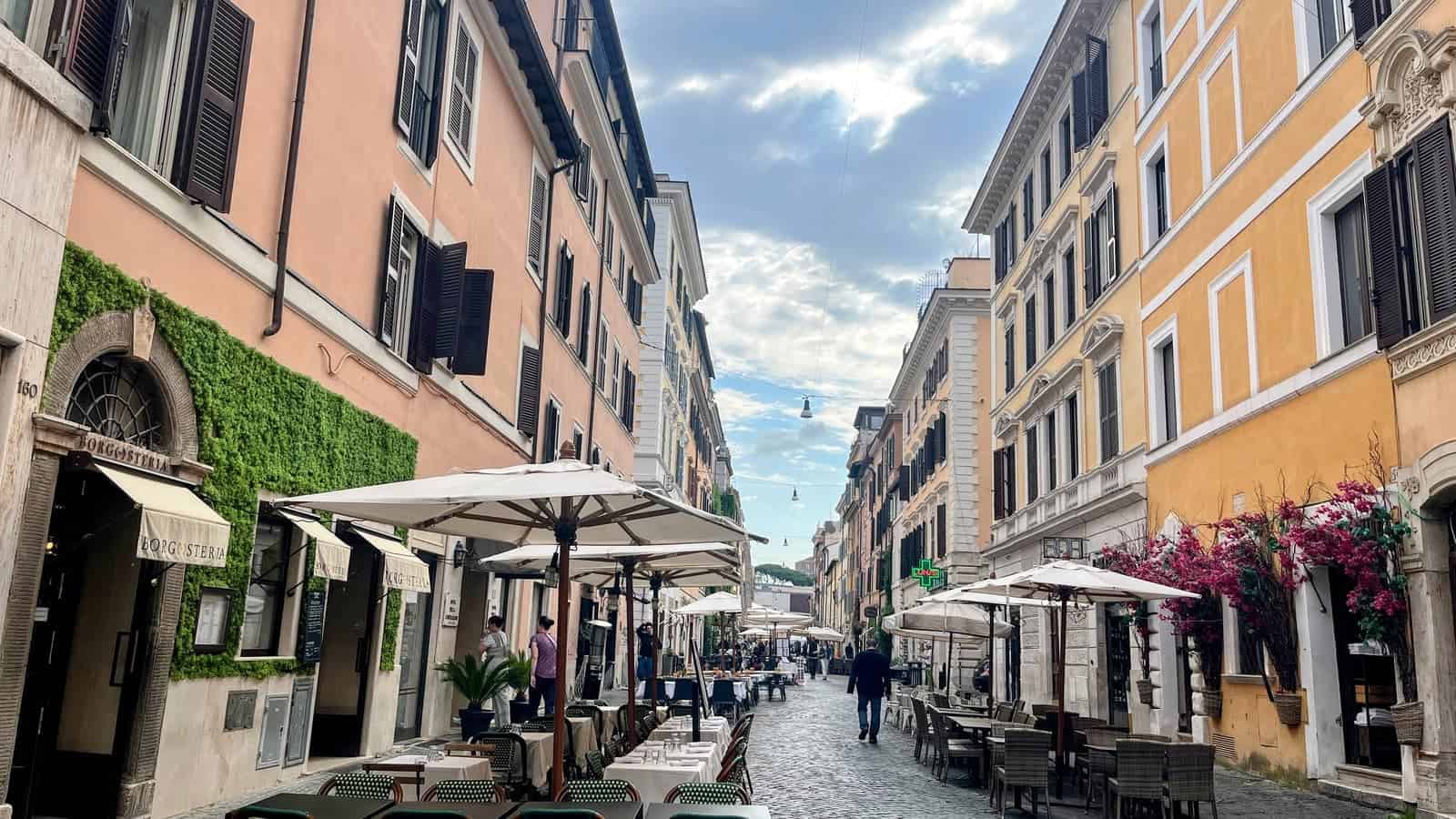 Cobblestone street in Rome lined with outdoor restaurant patios and cream colored umbrellas between historic buildings. Pink flowers and dramatic cloudy skies frame the lively dining area.