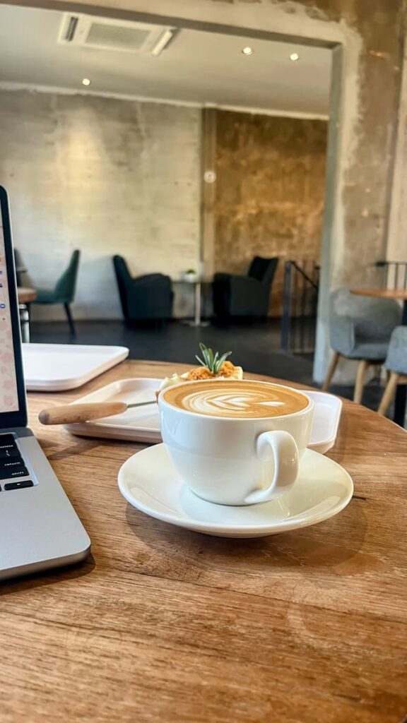 Latte with leaf shaped foam art served on a wooden table beside an open laptop in a modern café. Soft lighting and blurred lounge chairs create a quiet workspace atmosphere.