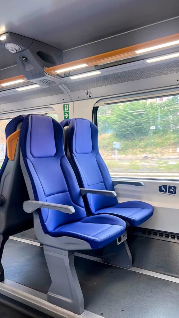 Two blue passenger seats beside a large train window on a modern train in Italy. Greenery and train tracks are visible outside the window during daytime travel.