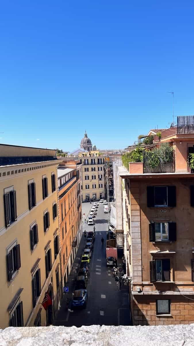 Narrow street lined with tall apartment buildings in Rome under a bright blue sky. Cars are parked along the cobblestone road and a domed church rises in the distance above the city skyline.