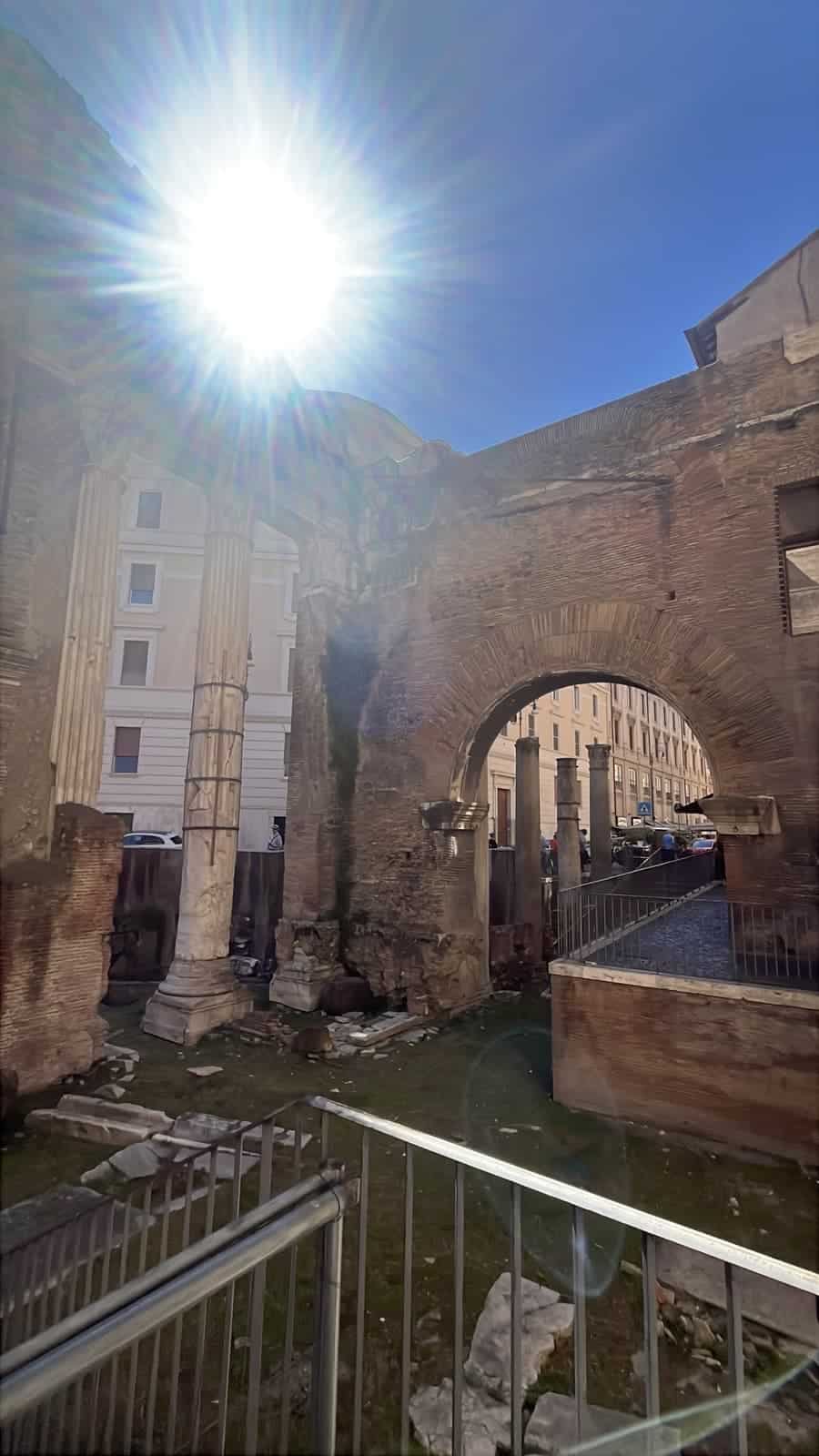 Ancient brick walls, marble columns, and a large stone arch frame an archaeological site in central Rome. Bright sunlight streams into the scene, highlighting the textures of the centuries-old ruins.