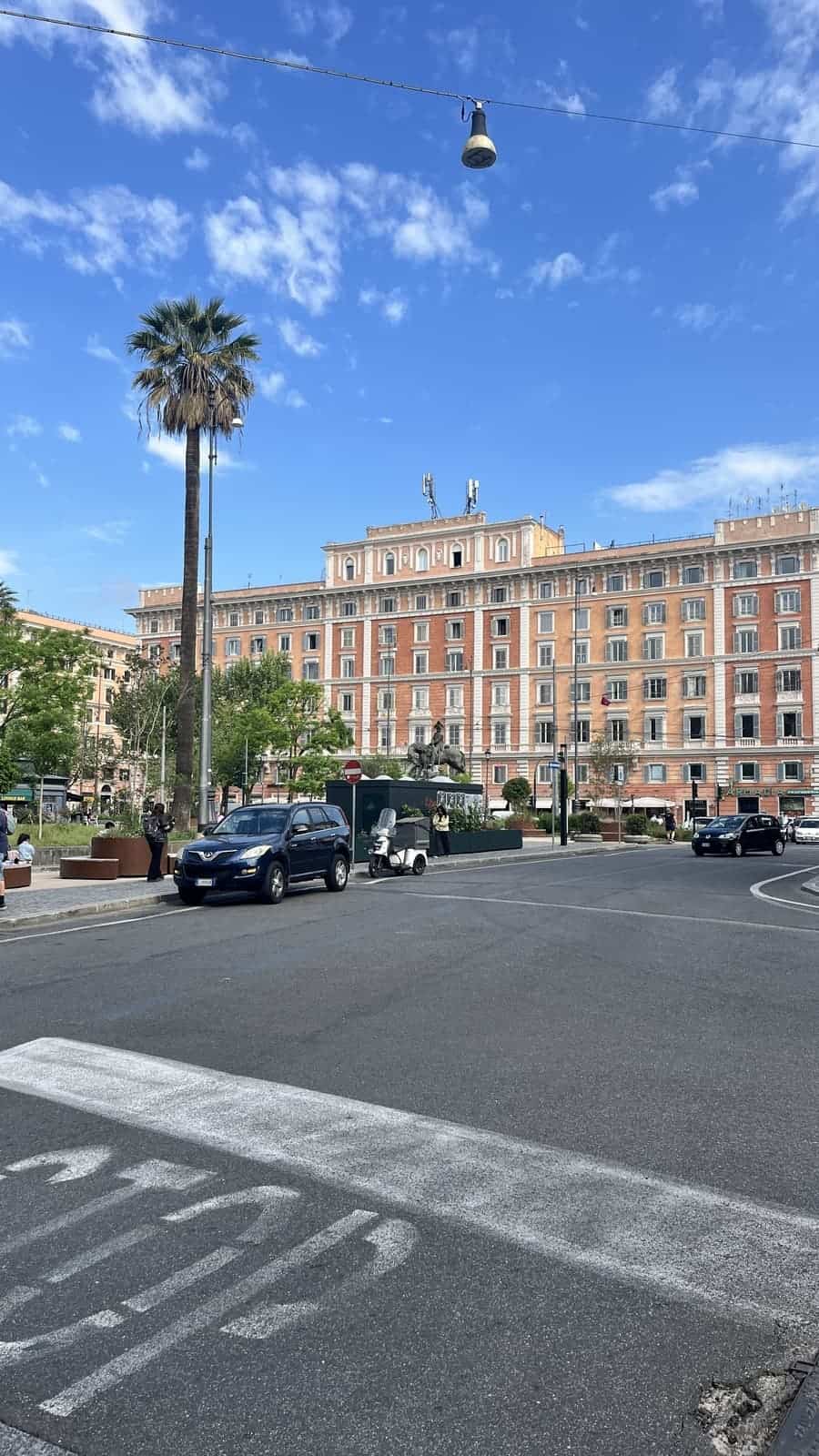 A broad Roman intersection features a tall palm tree, historic apartment buildings, and light traffic under a bright blue sky with scattered clouds. Cars, scooters, and pedestrians move through the neighborhood, highlighting everyday life in the city.
