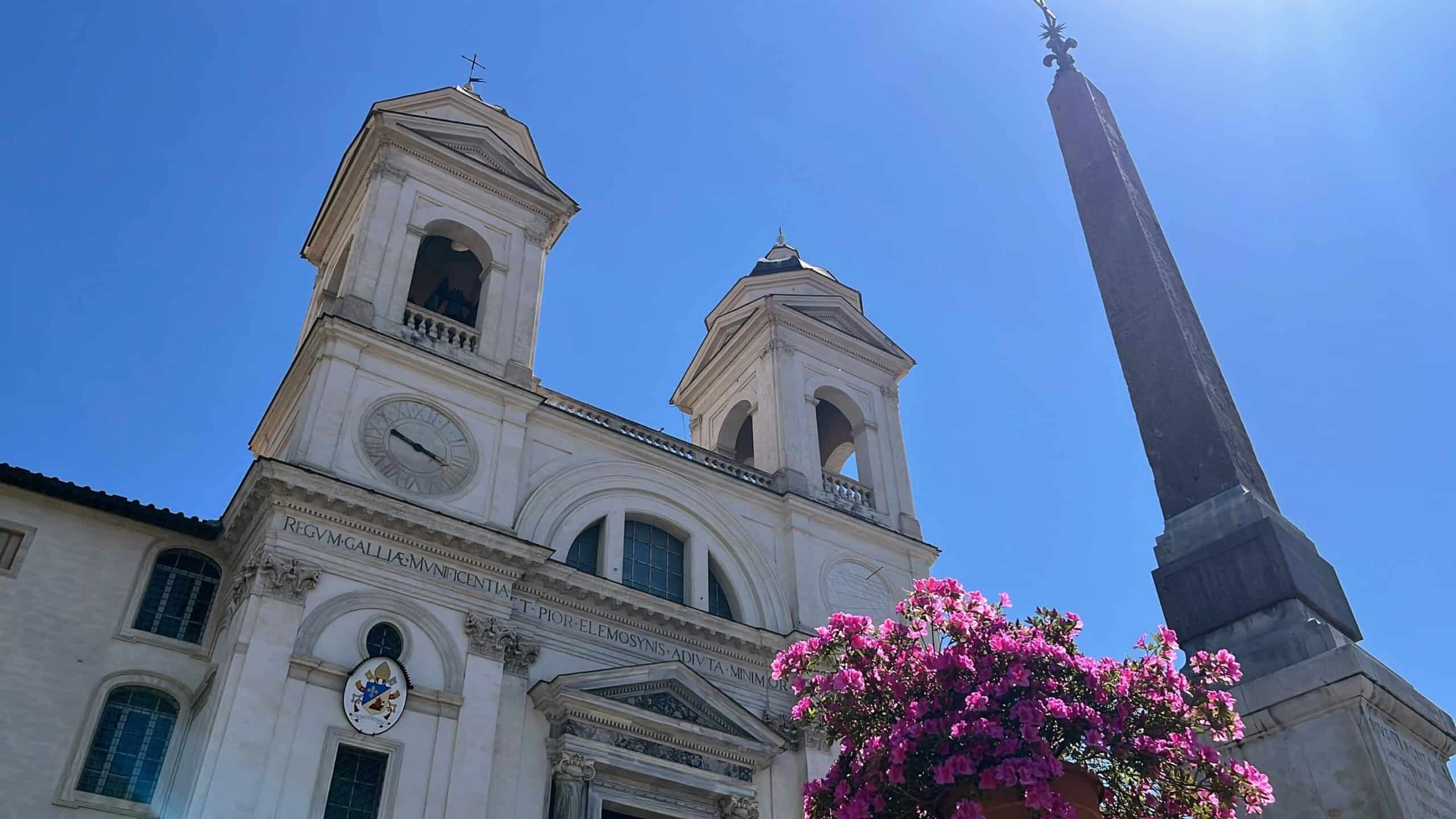 A low-angle view of Trinità dei Monti church and the Egyptian obelisk at the top of the Spanish Steps under a cloudless blue sky. Pink azaleas bloom in the foreground, adding color to the historic landmark.