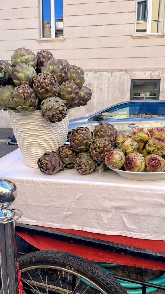 Fresh purple-green artichokes are stacked in a large white basket and arranged on platters at an outdoor market stall. The display sits on a white tablecloth with a parked car and city buildings in the background.