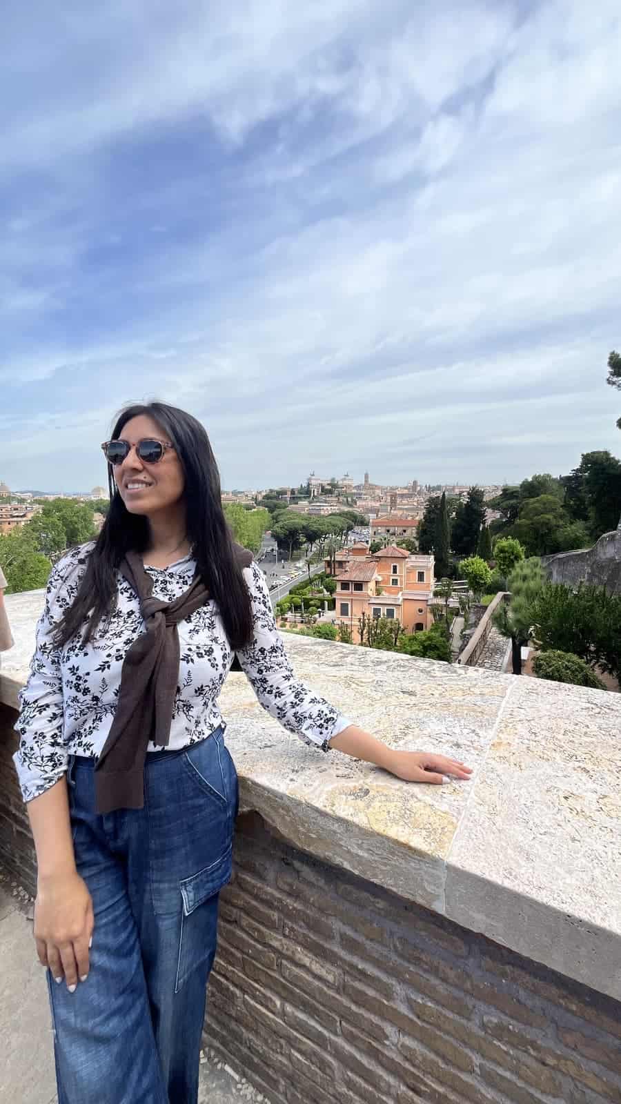 A smiling person wearing sunglasses, a floral blouse, and a denim skirt stands beside a stone terrace overlooking the rooftops and gardens of Rome. The elevated viewpoint offers a sweeping cityscape beneath a dramatic, cloud-filled sky.