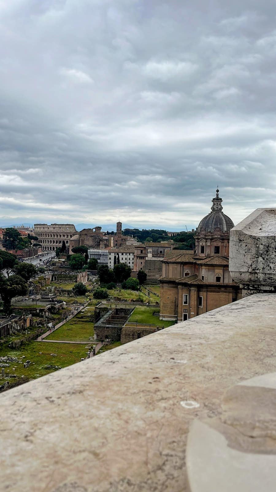 An elevated view overlooks the Roman Forum with the Colosseum visible in the distance beneath a dramatic overcast sky. Ancient ruins, historic churches, and archaeological remains stretch across the landscape, showcasing the heart of ancient Rome.