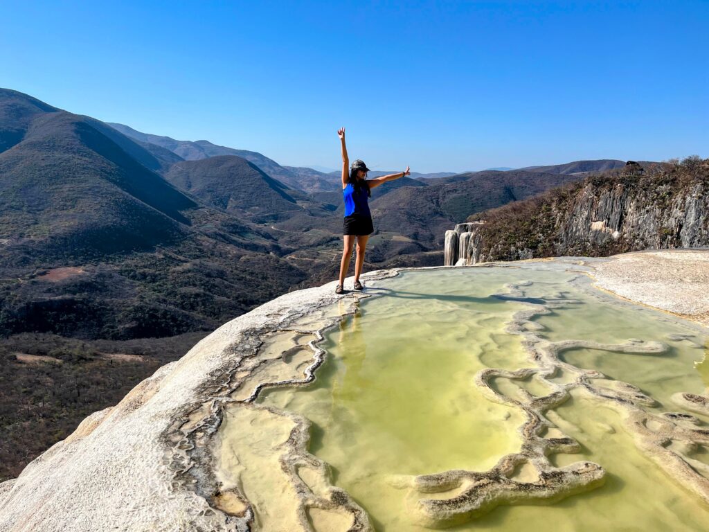 A traveler stands on the edge of the mineral pools at Hierve el Agua with arms raised, overlooking rugged mountains and the famous petrified waterfall in the distance. The turquoise mineral water contrasts with the dry landscape and clear blue sky, creating a dramatic panoramic view.