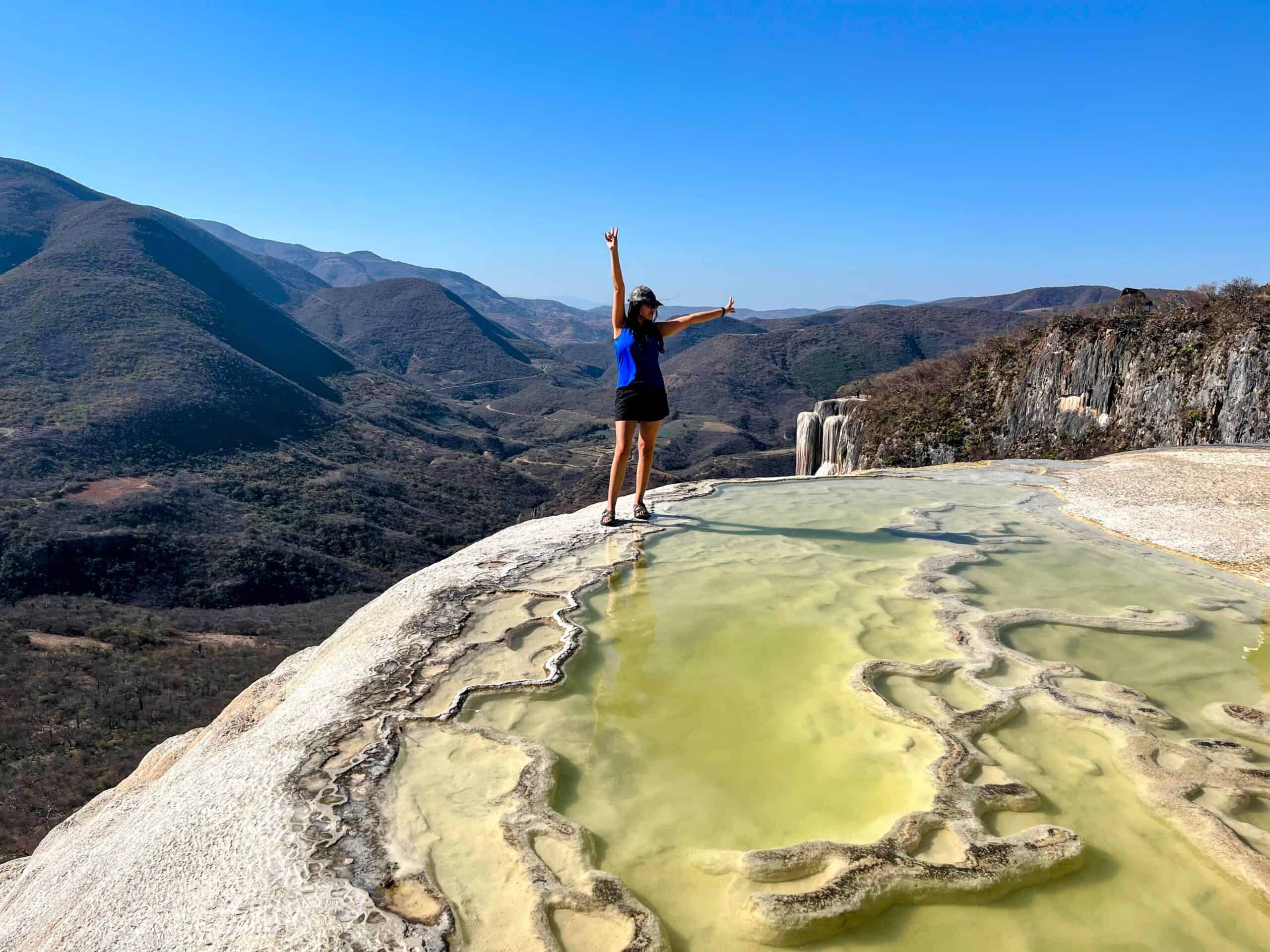 A traveler stands on the edge of the mineral pools at Hierve el Agua with arms raised, overlooking rugged mountains and the famous petrified waterfall in the distance. The turquoise mineral water contrasts with the dry landscape and clear blue sky, creating a dramatic panoramic view.