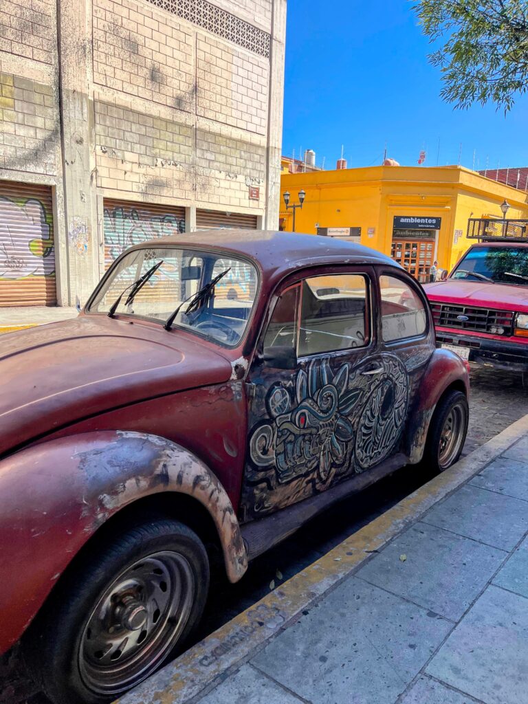 A weathered burgundy Volkswagen Beetle with a hand-painted black-and-white mural covering its door is parked along a sunny street in Oaxaca. The surrounding colorful buildings and classic architecture highlight the city's artistic character.