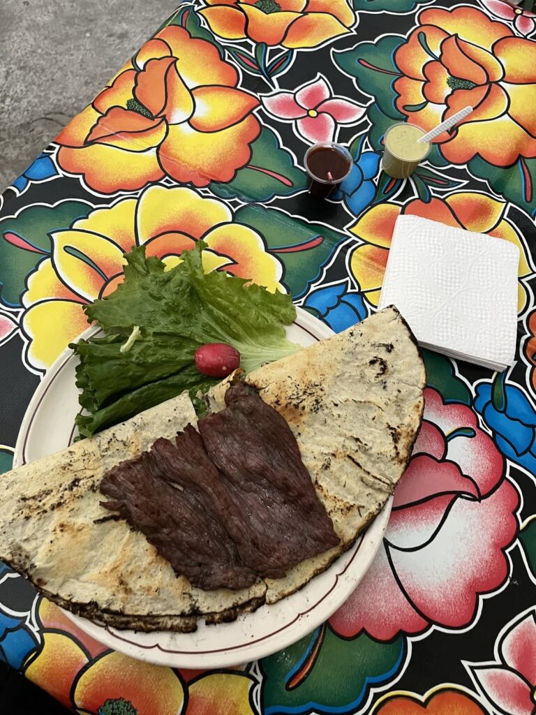 A plate of grilled meat served on a large grilled cactus paddle sits beside fresh lettuce, a radish, and small cups of salsa. The meal is placed on a vibrant floral tablecloth with colorful Mexican-inspired patterns.