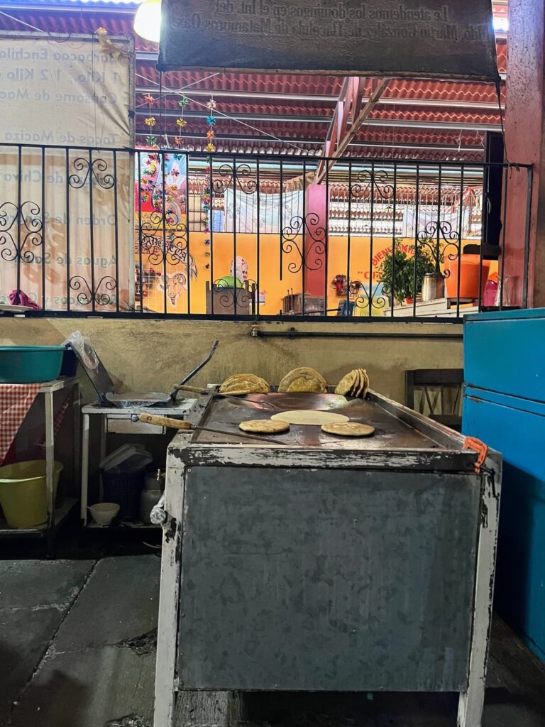 Fresh tortillas cook on a large flat-top griddle inside a bustling market food stall. Several finished tortillas are stacked nearby while the colorful kitchen and seating area are visible in the background.