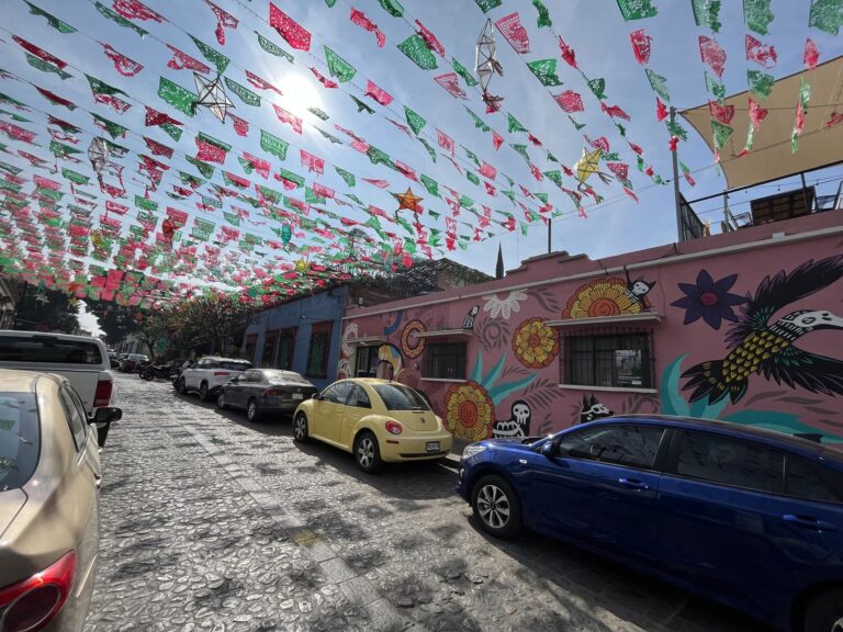 Rows of green, white, and red papel picado and decorative star lanterns hang above a cobblestone street lined with colorful murals and parked cars. Sunlight filters through the decorations, creating a festive streetscape inspired by Mexican celebrations.