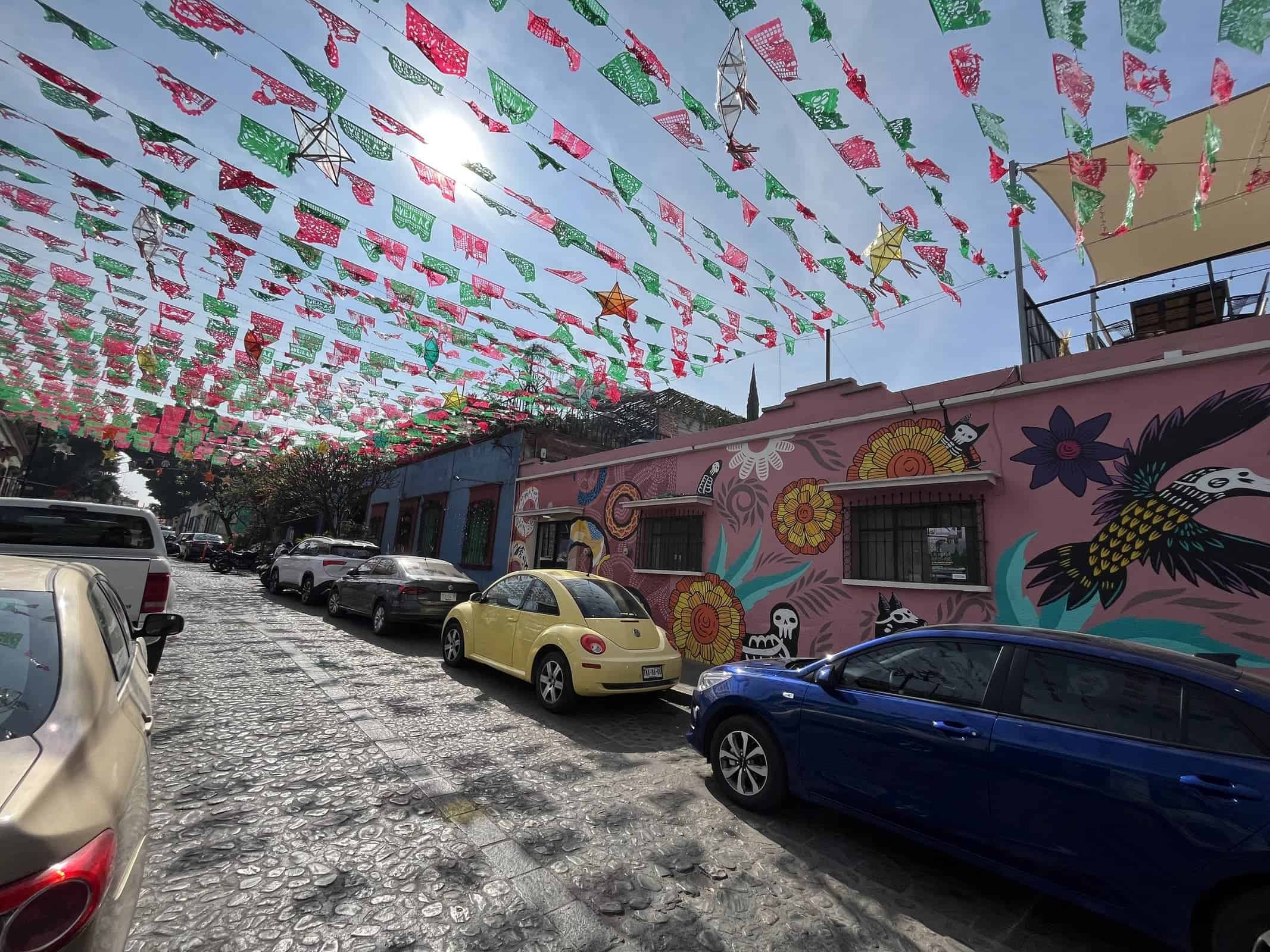 Rows of green, white, and red papel picado and decorative star lanterns hang above a cobblestone street lined with colorful murals and parked cars. Sunlight filters through the decorations, creating a festive streetscape inspired by Mexican celebrations.