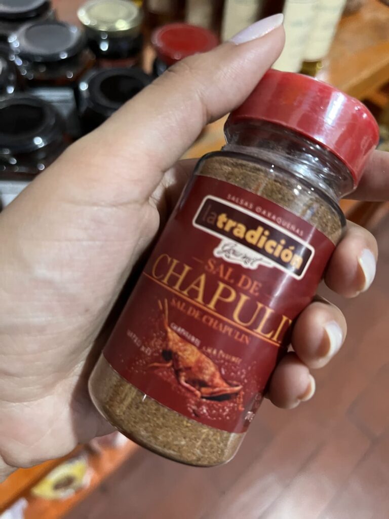 A hand holds a small jar labeled "La Tradición Sal de Chapulín," a seasoning made with ground grasshoppers. Additional spice jars are softly blurred in the background on a wooden display shelf.