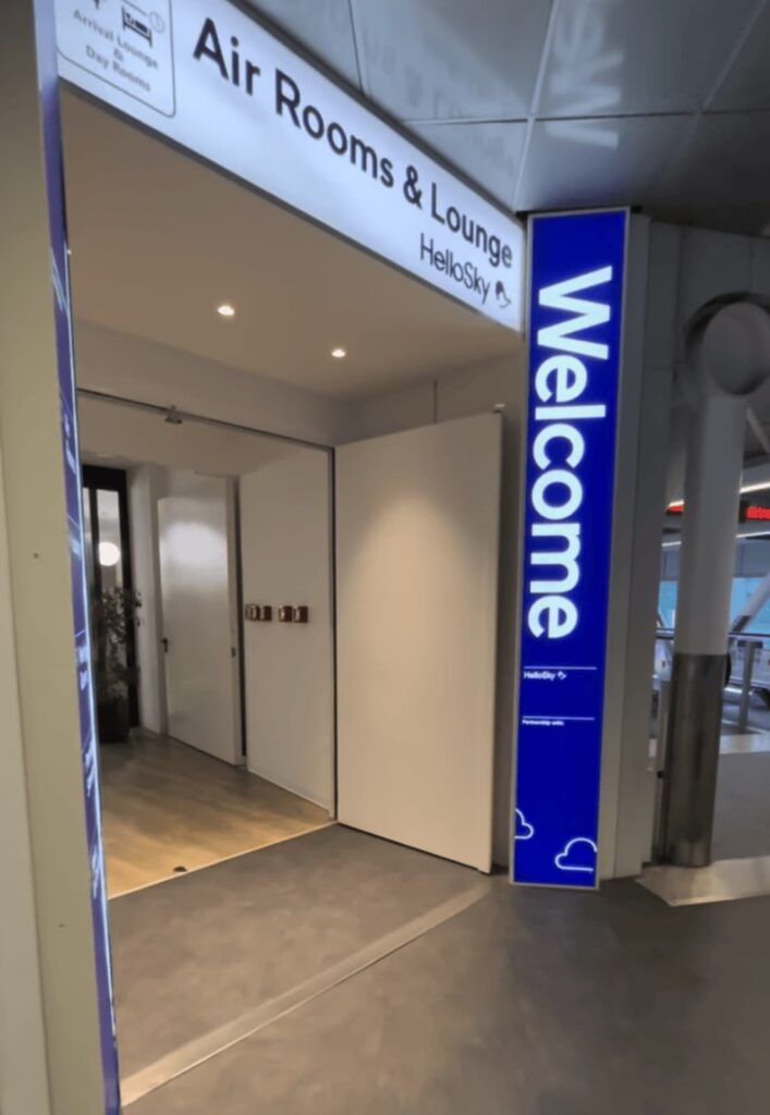 Entrance to the “Air Rooms & Lounge” by HelloSky inside an airport. A tall illuminated blue sign beside the doorway reads “Welcome.”