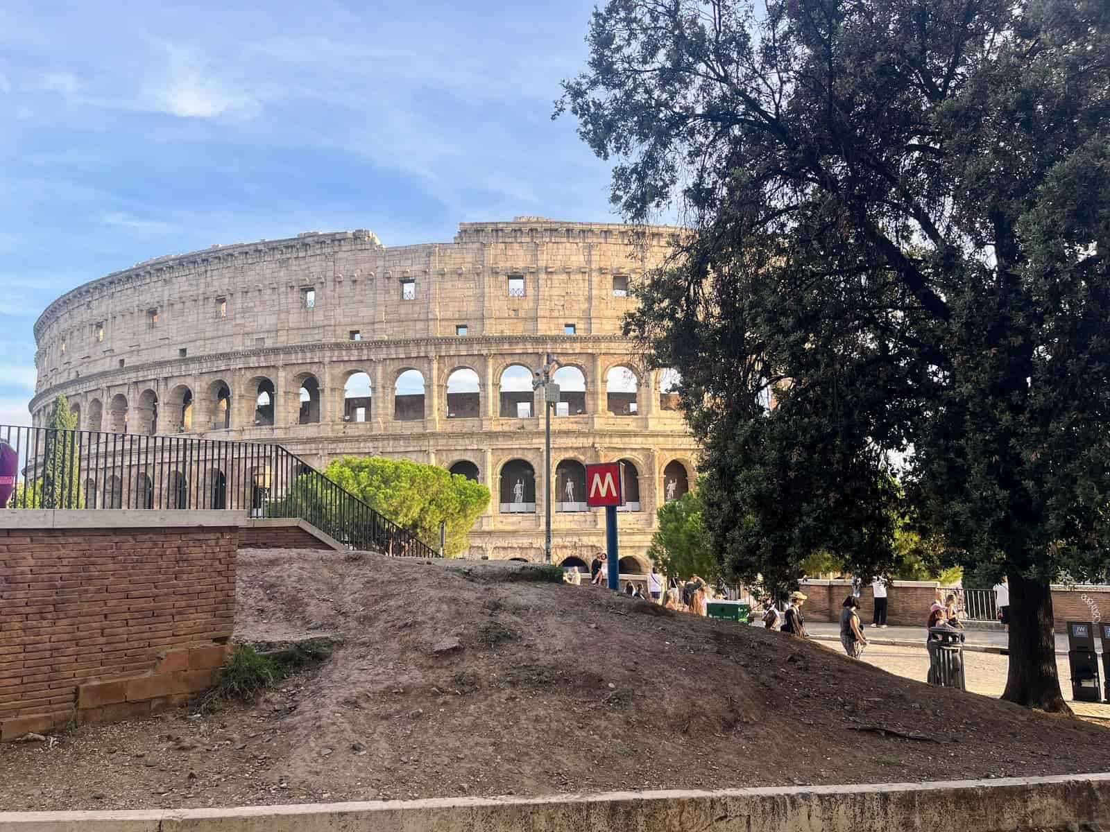The Colosseum in Rome seen in warm afternoon light with a red Metro sign in front. Trees and pedestrians surround the ancient amphitheater under a blue sky.